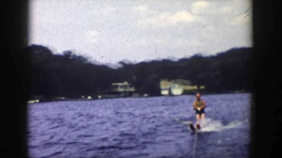 1958 Wisconsin 8mm frame of a young man water skiing on a lake, with trees, lakeside buildings and cloudy sky in the distance.