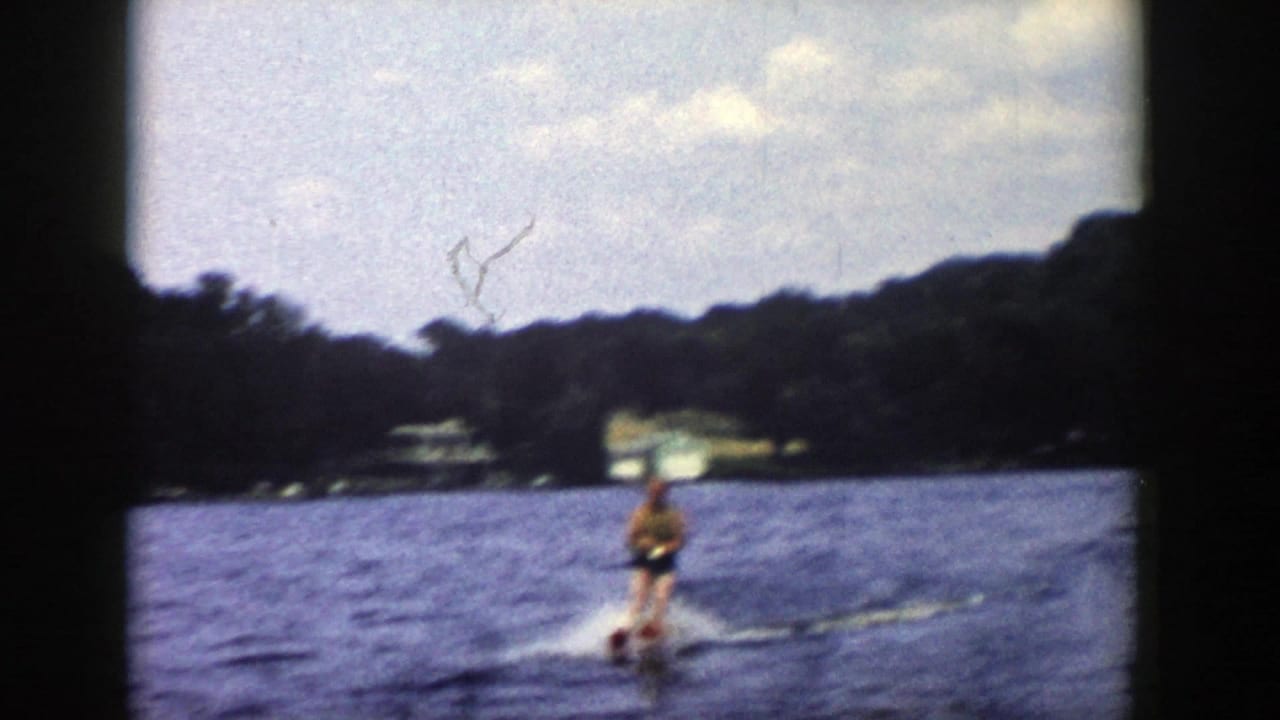 The skier remains upright on red skis in a distant 1958 Wisconsin lake scene, framed by shoreline trees and lakeside structures.