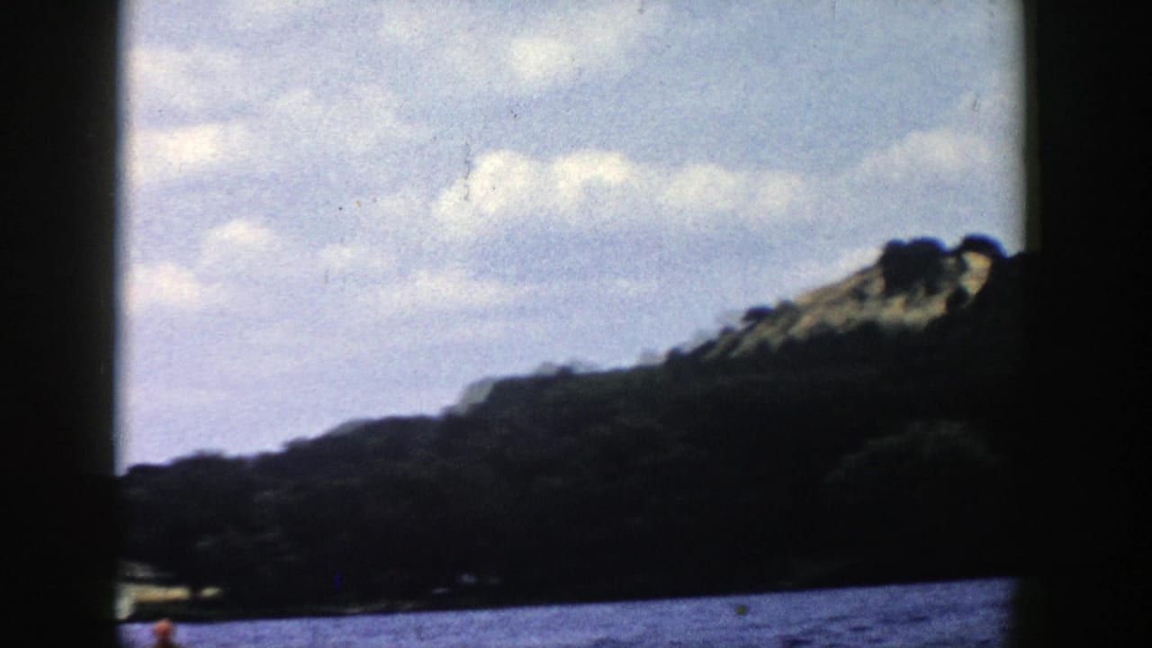 Wide 8mm home-movie view of a Wisconsin lake shoreline and cloudy sky, with the water skier partly framed near the bottom edge.