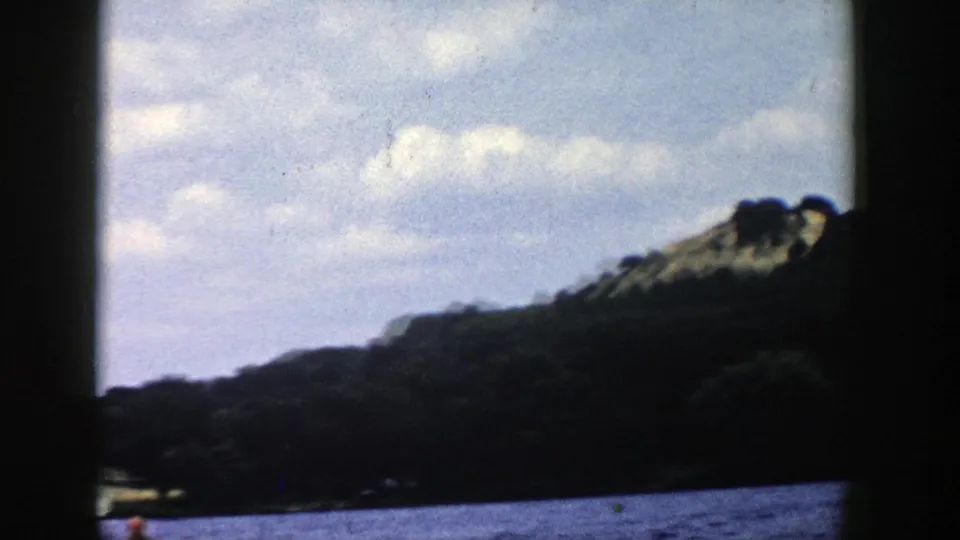 Wide 8mm home-movie view of a Wisconsin lake shoreline and cloudy sky, with the water skier partly framed near the bottom edge.