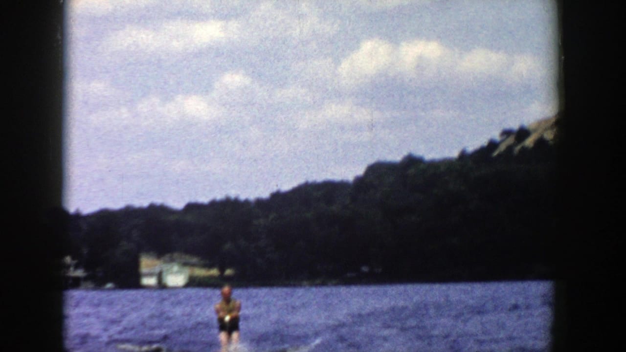 A distant skier crosses a Wisconsin lake in this 1958 8mm frame, with wooded shore, a pale building and soft clouds in view.