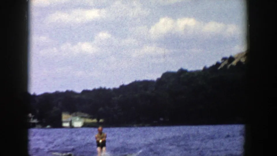 A distant skier crosses a Wisconsin lake in this 1958 8mm frame, with wooded shore, a pale building and soft clouds in view.