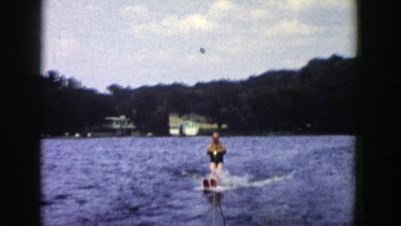 Water skier approaches across a lake with a tow rope and distant tree-lined shore.