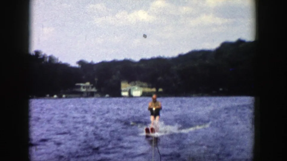 1958 Wisconsin home-movie frame showing a water skier in a yellow top and dark shorts moving across a lake near distant shore houses.