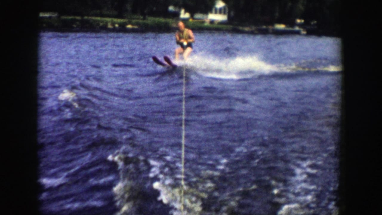 A 1958 Wisconsin home-movie frame captures the skier cutting across the wake, with water spray, tow rope, and lakeside buildings visible.