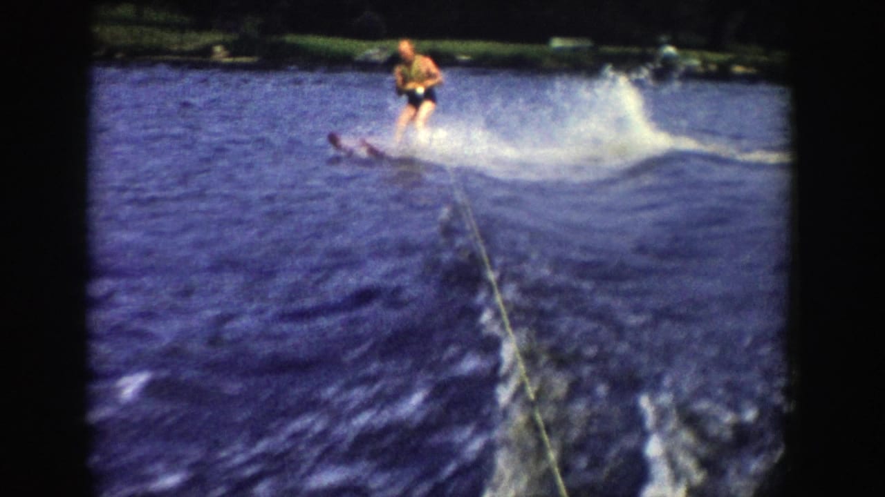 The skier leans through a turn on the Wisconsin lake, with a broad spray, tow rope, wake lines, and wooded shoreline behind.