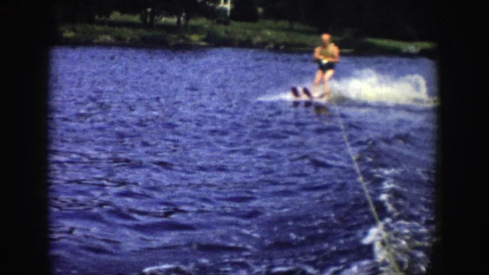 In this 1958 Wisconsin 8mm frame, a man water skis across a lake behind the filming boat, with spray, tow rope, and trees in the distance.