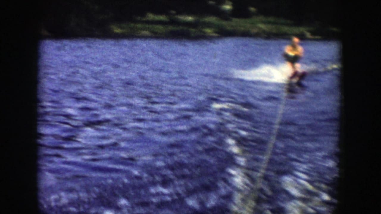 A 1958 Wisconsin home-movie frame shows the skier farther right on the lake, with the tow rope, wake, and blue water dominating the view.