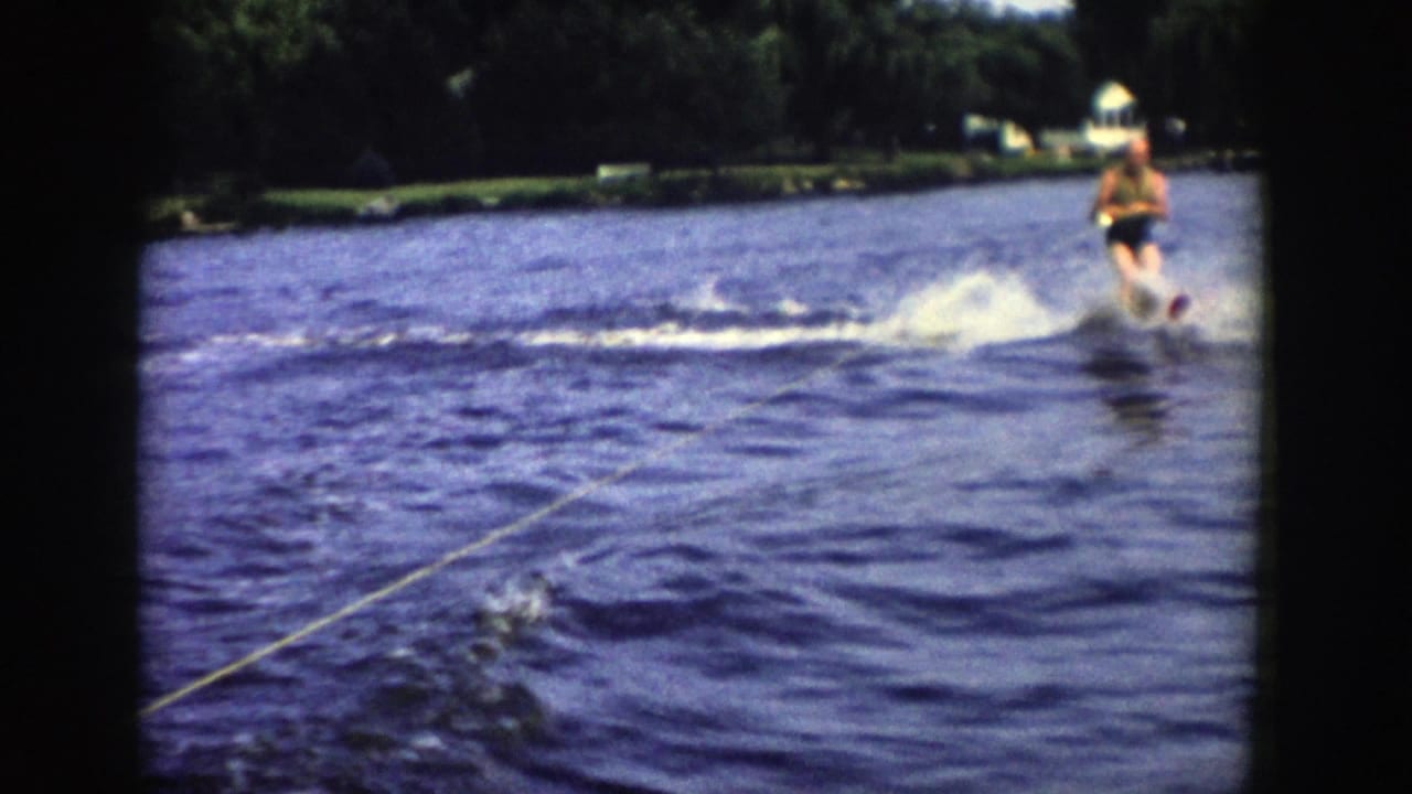 In 1958 Wisconsin home-movie footage, a water skier glides across a lake behind a tow rope, with shoreline trees in the distance.