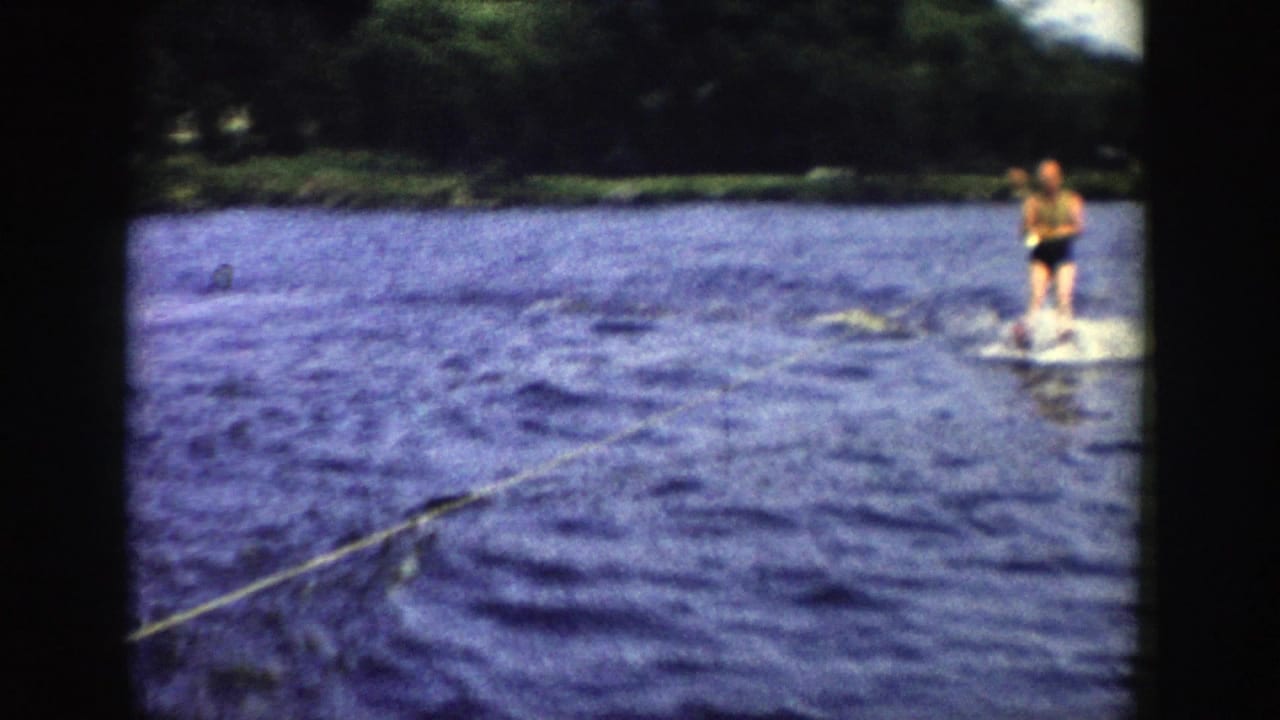 A water skier continues across a Wisconsin lake, framed by dark 8mm borders and a wooded shoreline under bright daylight.