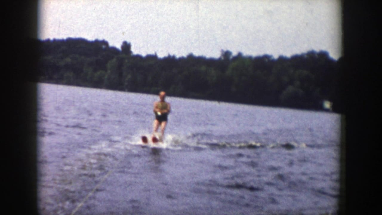 The skier stands upright on water skis in mid-lake, with a dark tree line and soft 8mm color in the background.