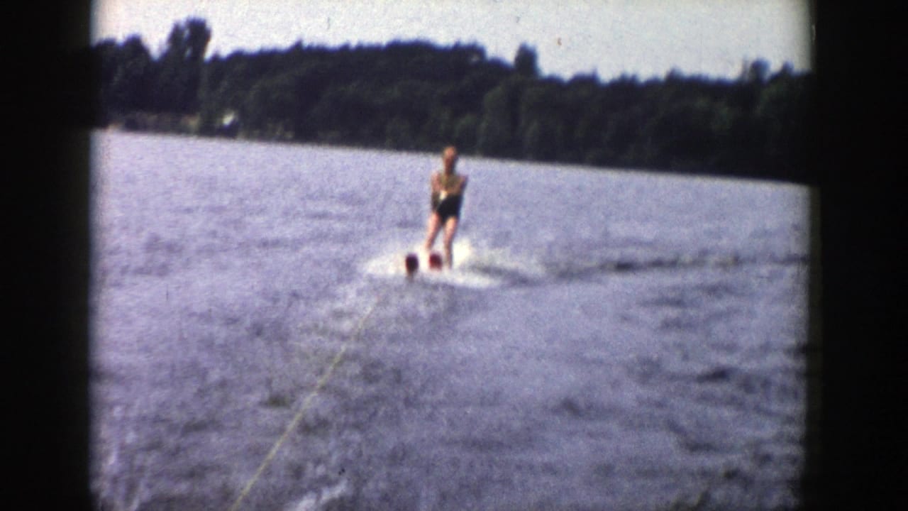 A Wisconsin lake water skier approaches along the tow line, throwing light spray against a wooded shoreline backdrop.