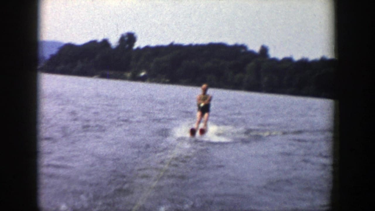 A man water skis on a lake while holding a tow rope, with shoreline and hills behind him.