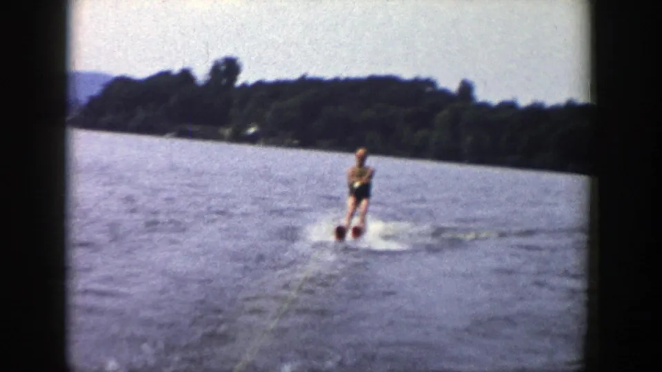 In 1958 Wisconsin home-movie footage, a man on water skis glides across a lake while holding a tow rope near a wooded shore.