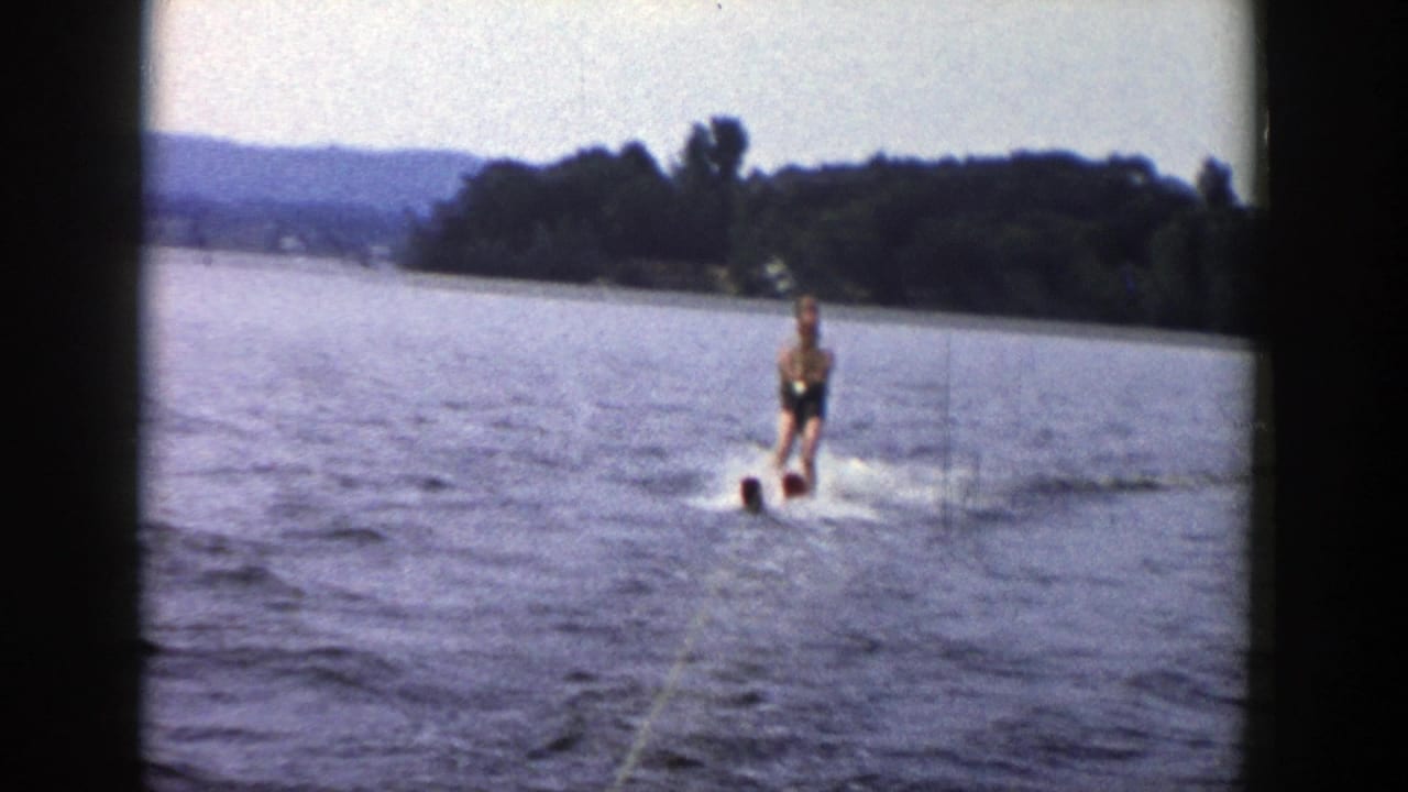A distant man water skis toward the camera on a Wisconsin lake, with tow rope, wake, shoreline, and hills visible in the 8mm frame.