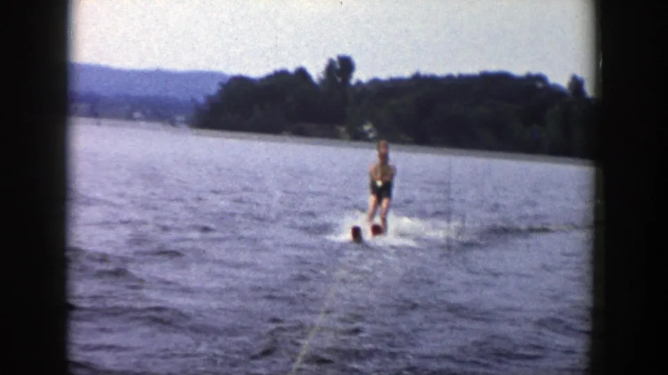 A distant man water skis toward the camera on a Wisconsin lake, with tow rope, wake, shoreline, and hills visible in the 8mm frame.