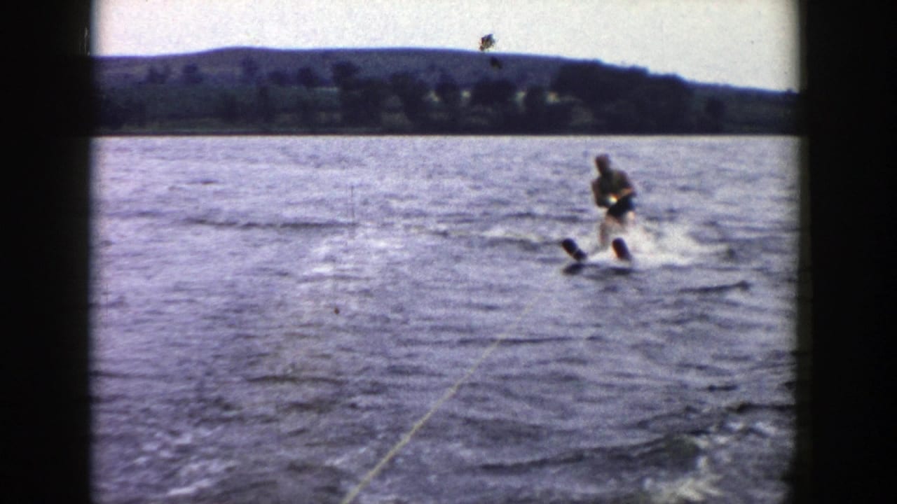 A man water skis across a Wisconsin lake, leaning as he follows the tow rope with wooded shoreline and low hills beyond.