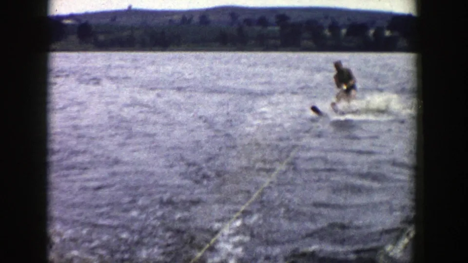 1958 Wisconsin 8mm frame showing a man water skiing behind a boat, with wake, spray, tow rope, and a tree-lined shore in the distance.