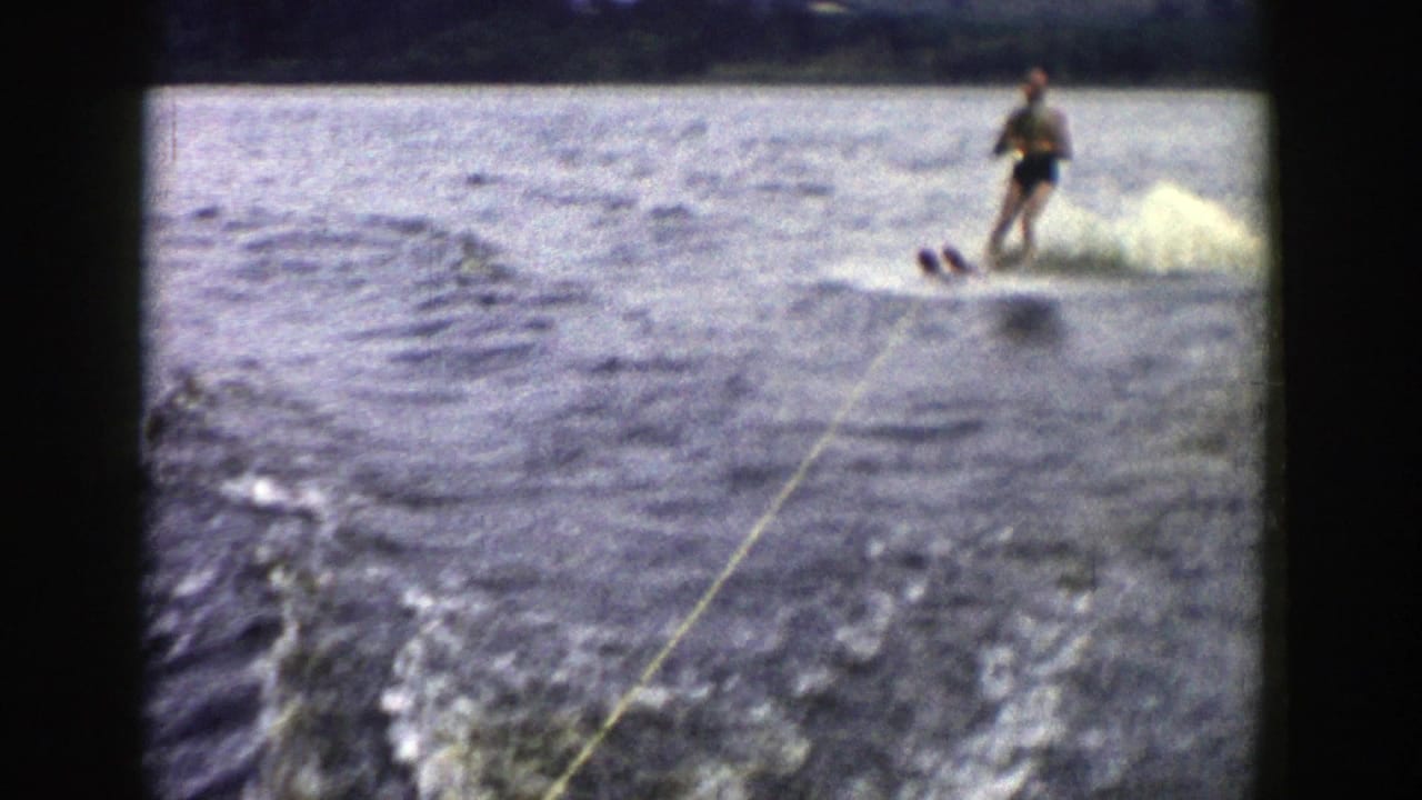 A man water skis over choppy lake water in 1958 Wisconsin home-movie footage, with tow rope, wake, spray, and shore visible beyond.