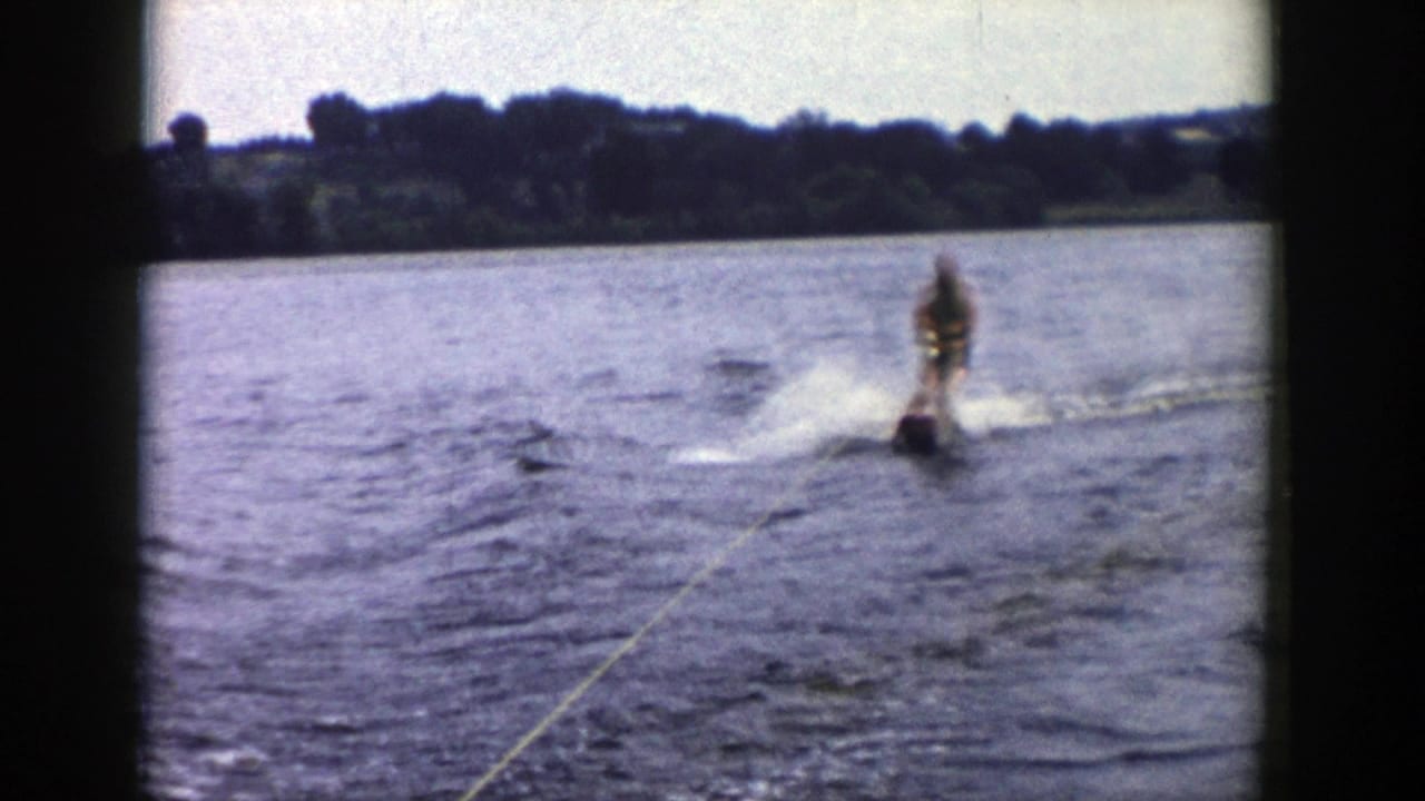 1958 Wisconsin lake scene with a male water skier approaching behind the tow rope, surrounded by wake spray and a dark shoreline.