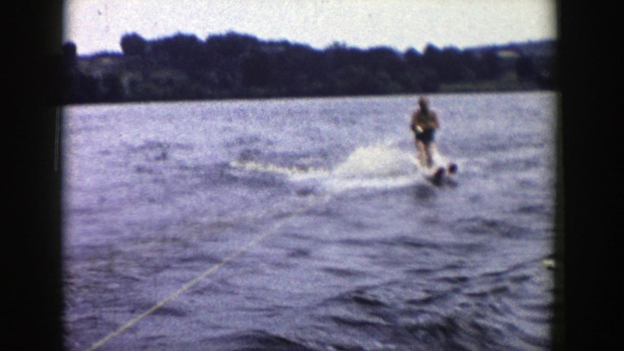 A 1958 Wisconsin 8mm frame shows the skier riding at frame right, with tow rope, wake, spray, open water, and tree-lined shore behind.