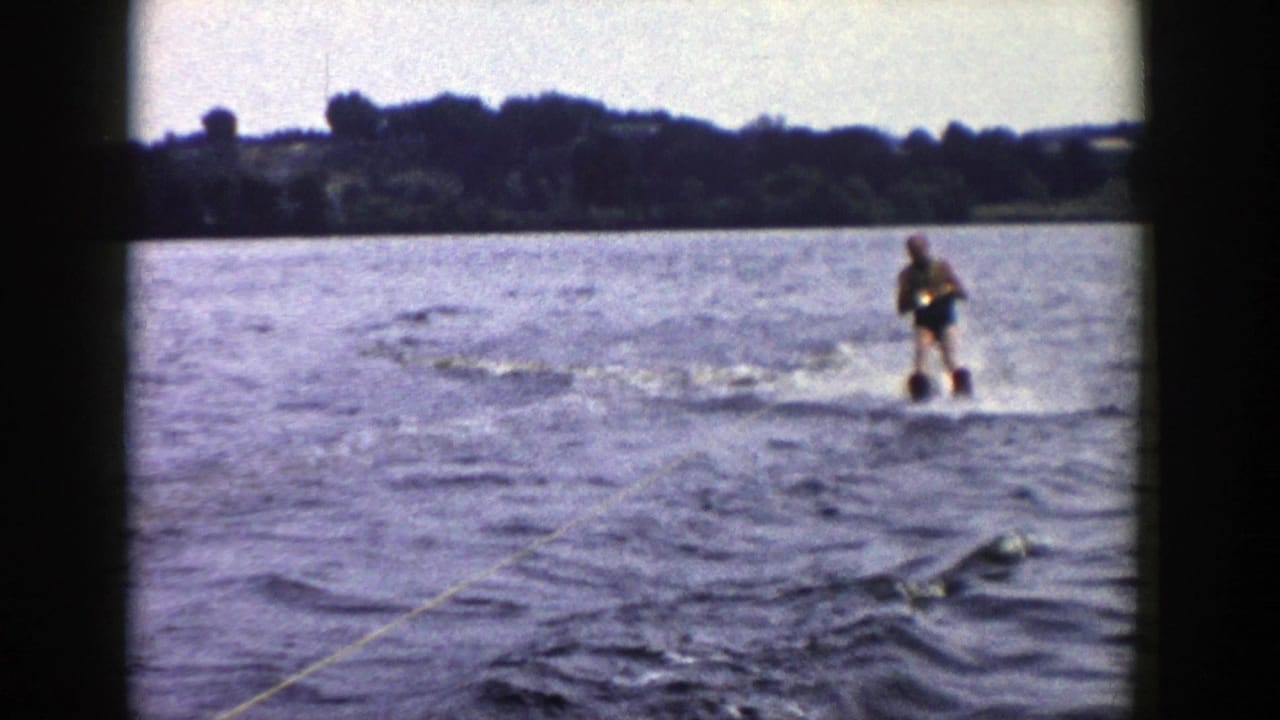 1958 Wisconsin 8mm frame showing a man in a dark swimsuit water skiing across a lake, with tow rope, choppy water, and trees in the distance.