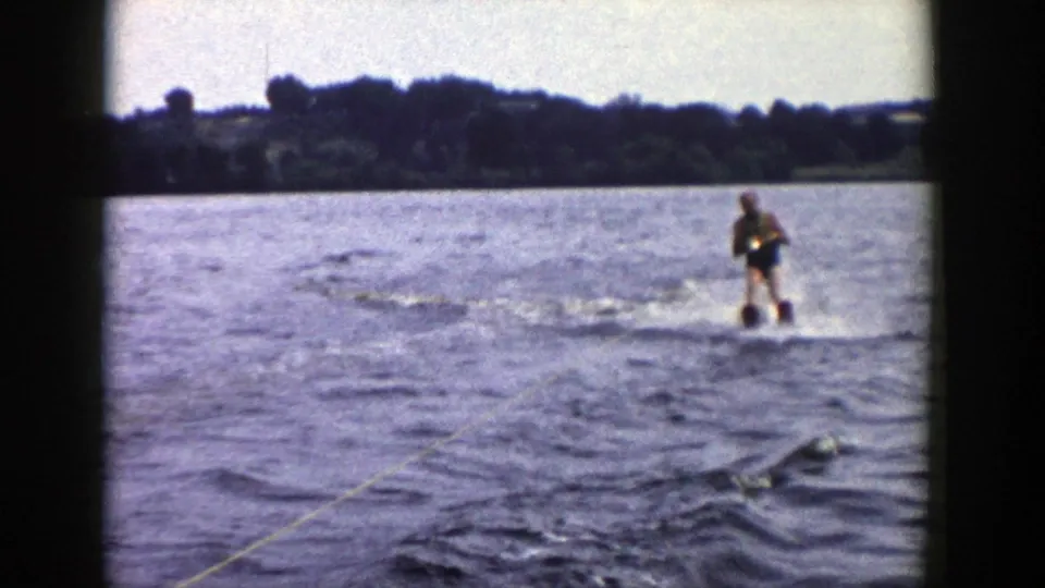 1958 Wisconsin 8mm frame showing a man in a dark swimsuit water skiing across a lake, with tow rope, choppy water, and trees in the distance.