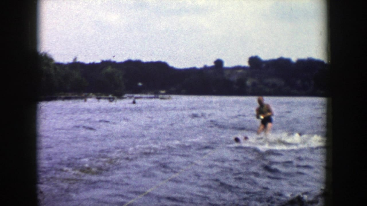 1958 Wisconsin home-movie frame with a man water skiing near distant swimmers or heads in the water, backed by a wooded lakeshore.