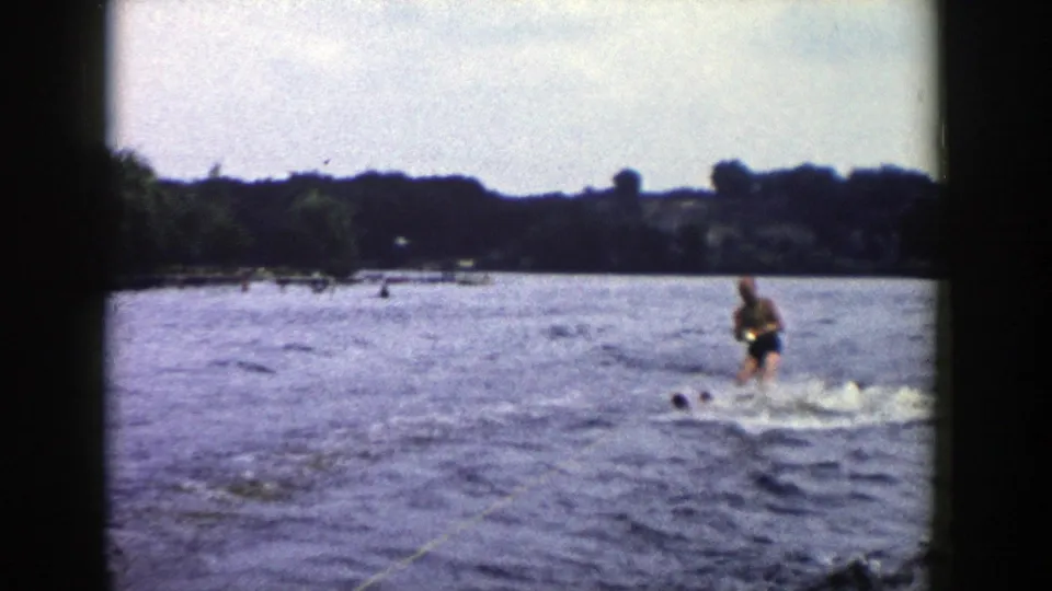1958 Wisconsin home-movie frame with a man water skiing near distant swimmers or heads in the water, backed by a wooded lakeshore.