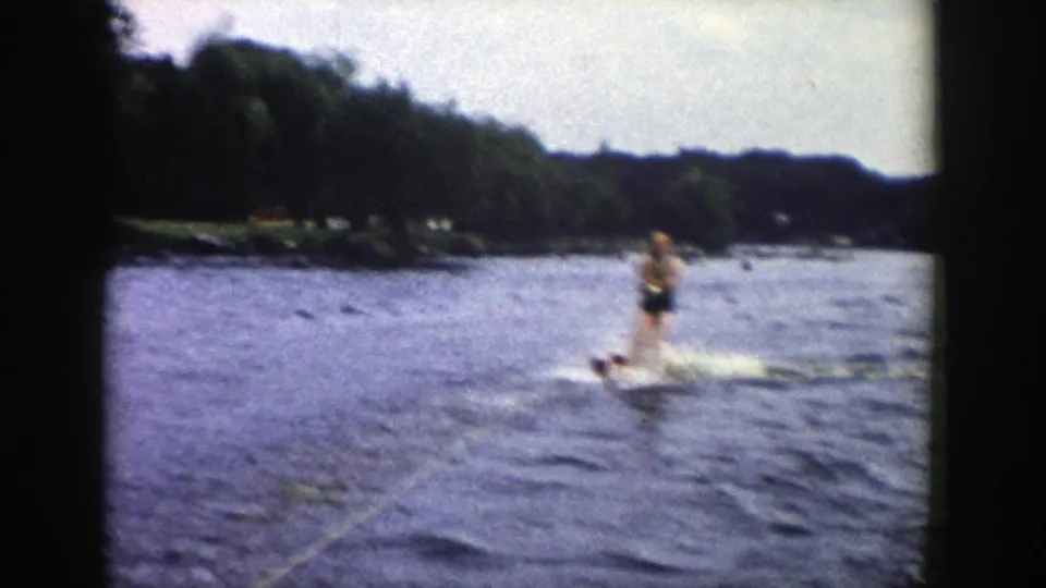 1958 Wisconsin 8mm frame showing a water skier approaching over choppy lake water, with a tow rope in the foreground and trees along the shore.