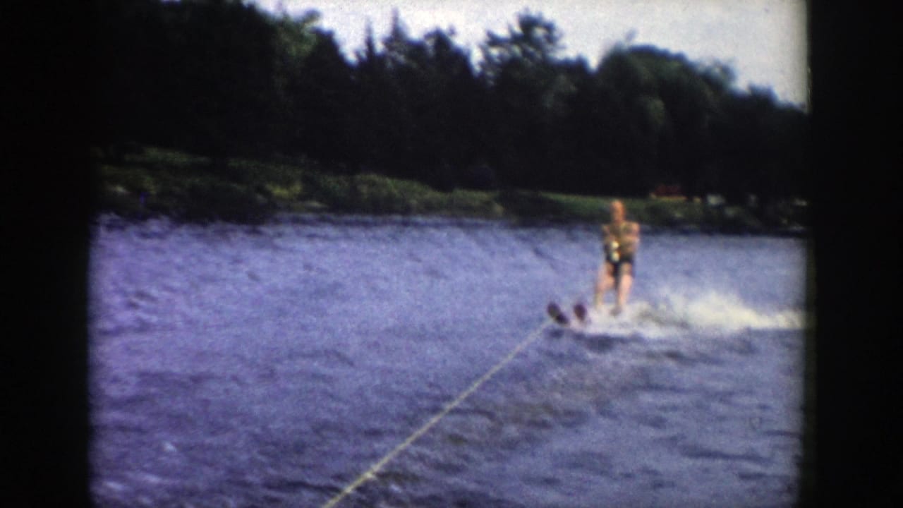 1958 Wisconsin home-movie frame with a man water skiing through spray, tow rope visible in front, and a tree-lined lakeshore in the background.