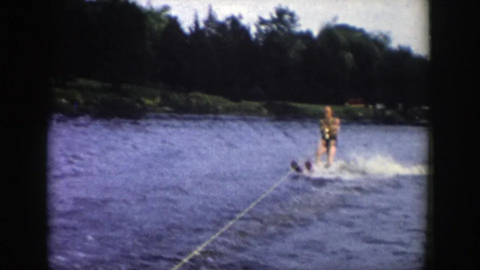 1958 Wisconsin home-movie frame with a man water skiing through spray, tow rope visible in front, and a tree-lined lakeshore in the background.