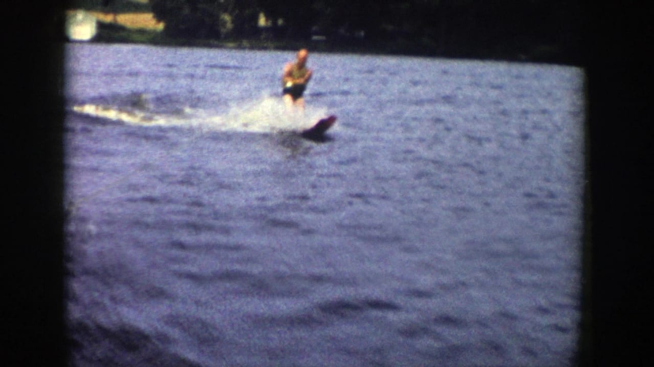 Man water skiing across a lake with spray behind him and trees on the far shore.