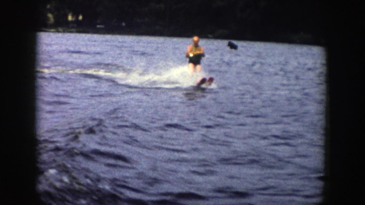 A man in a dark swimsuit skis toward the camera boat on a Wisconsin lake, with spray and a distant shoreline visible.