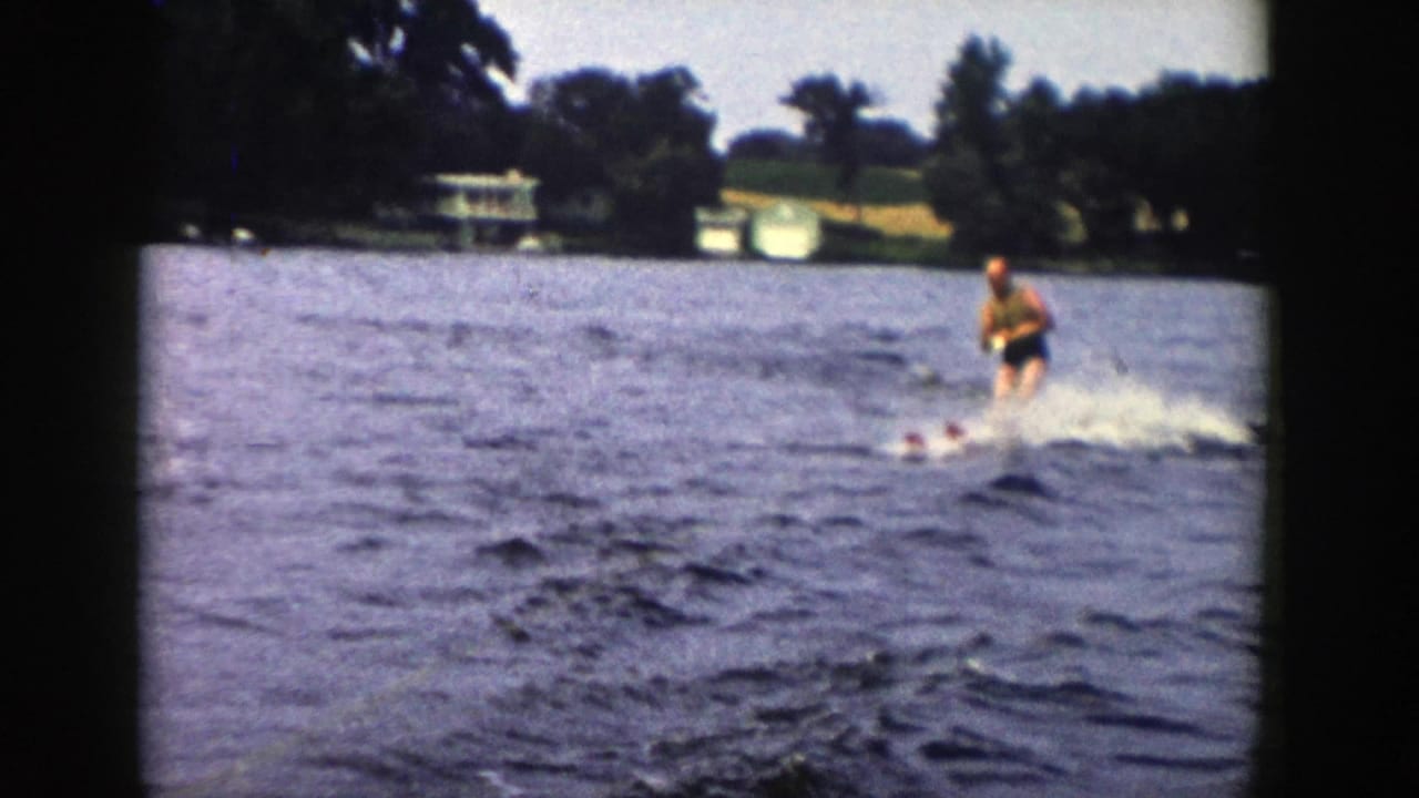 Man water skiing on a lake with shoreline trees and small structures in the distance.