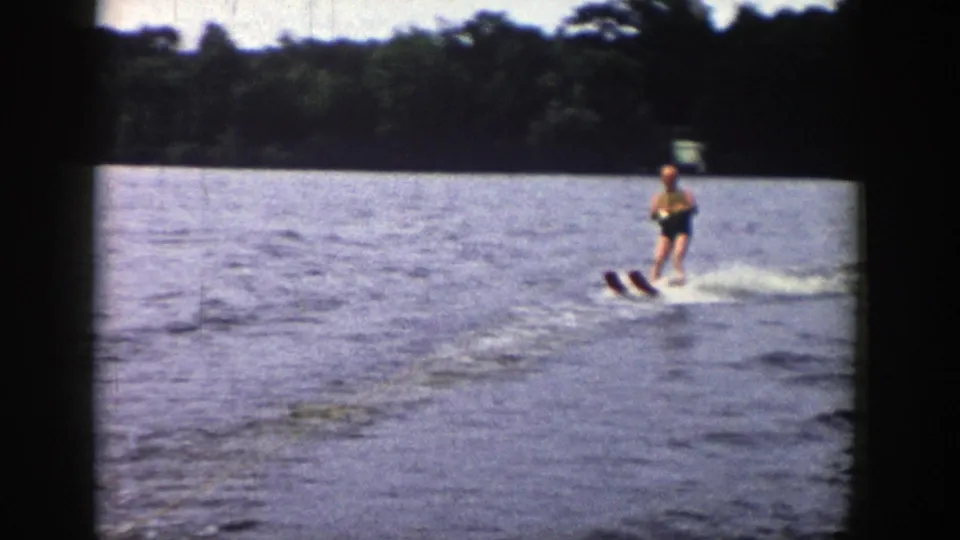 Young person water skiing on a Wisconsin lake in 1958, seen in soft 8mm color with wooded shoreline behind.