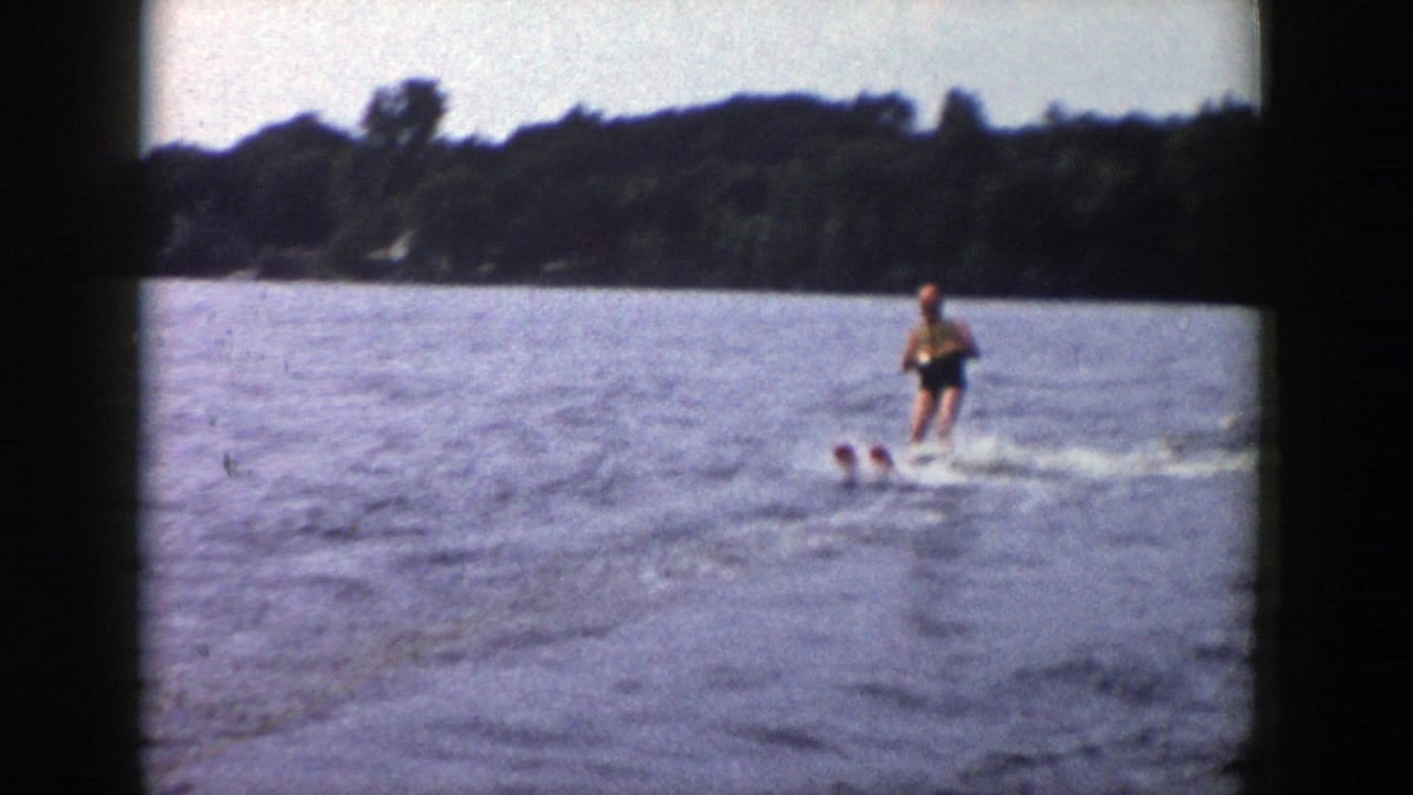 The skier continues across open water, with a dark wooded shore and hazy sky in the background.