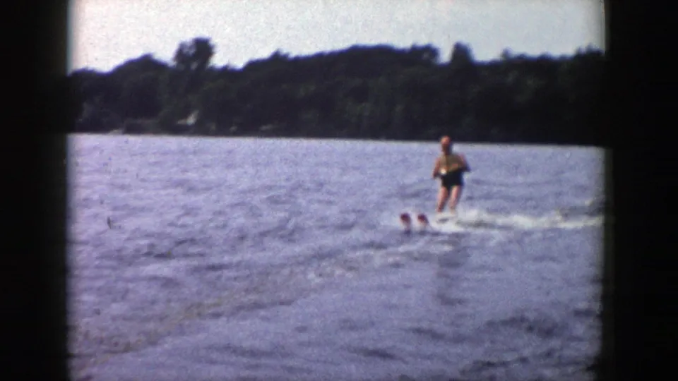 The skier continues across open water, with a dark wooded shore and hazy sky in the background.