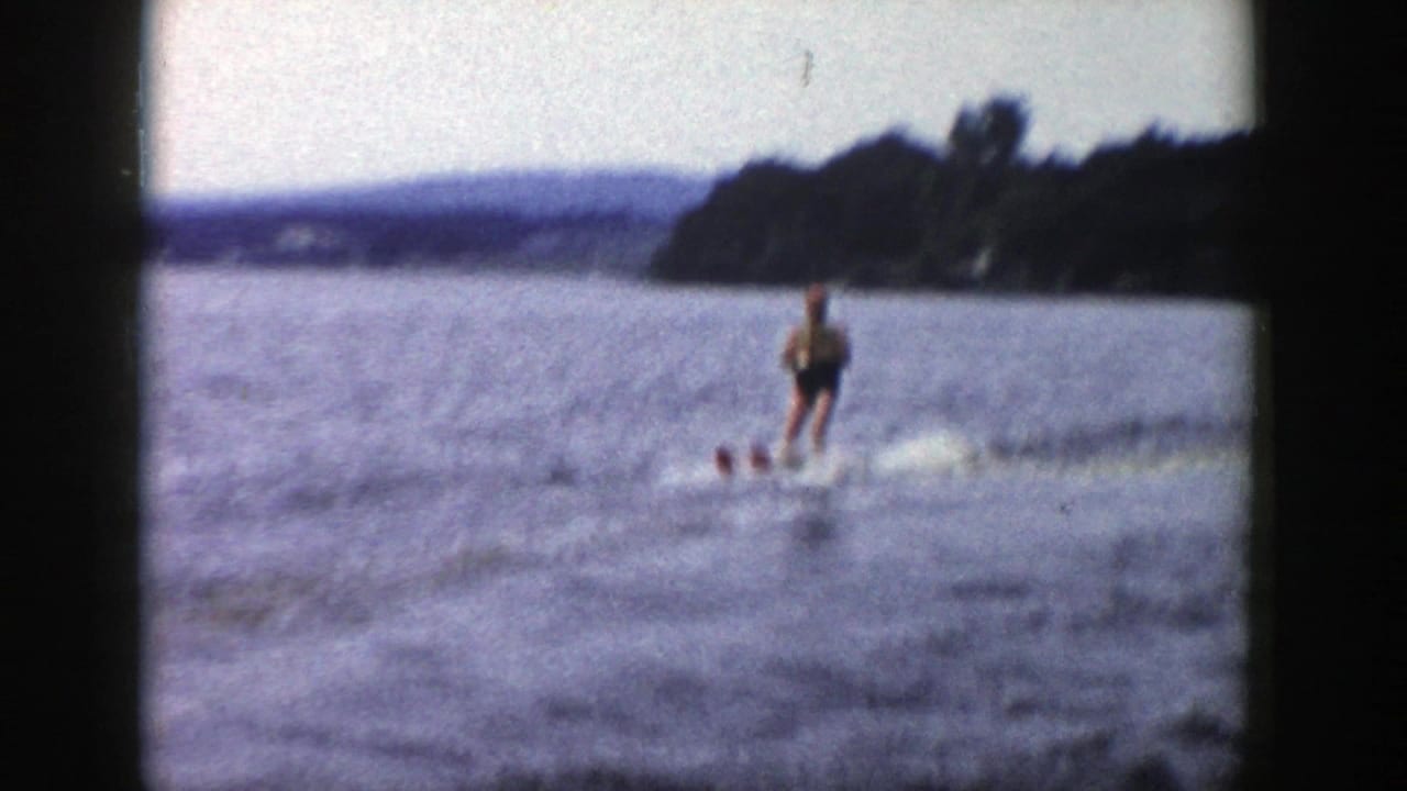 A soft, grainy 8mm frame shows the skier upright on the lake, with distant hills and trees beyond the water.
