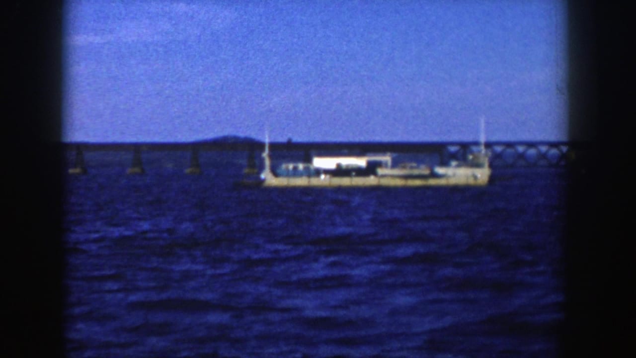1958 Wisconsin 8mm frame showing a white stationary vessel on rippled water below a long low bridge and distant shoreline.