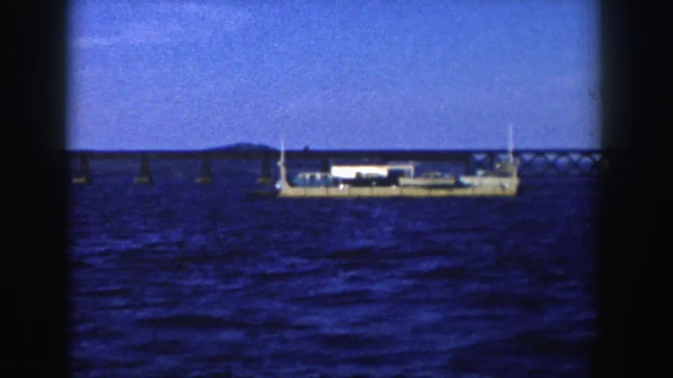 1958 Wisconsin 8mm frame showing a white stationary vessel on rippled water below a long low bridge and distant shoreline.