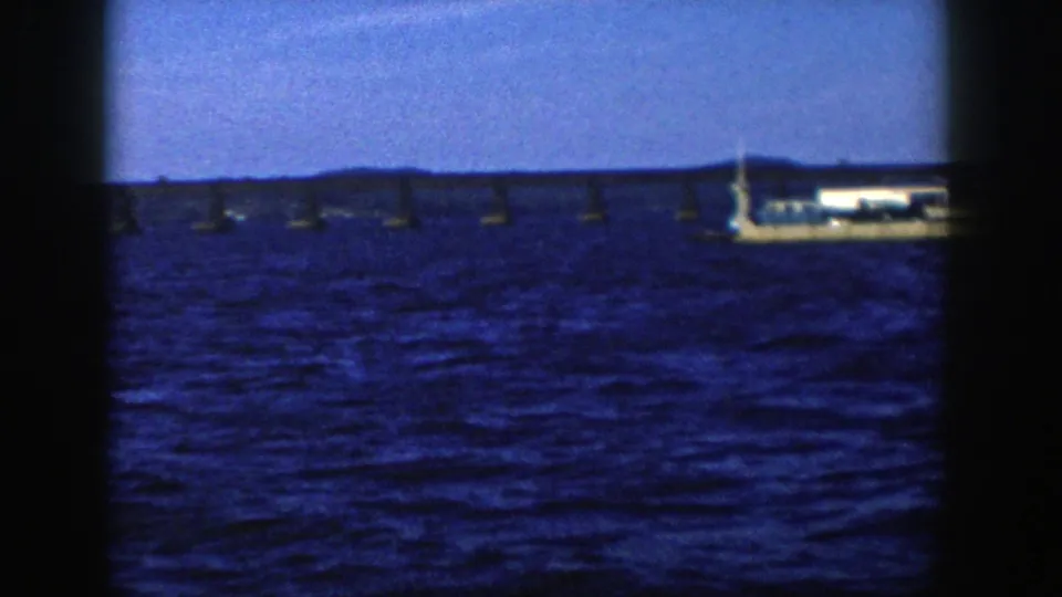 1958 Wisconsin 8mm frame with a white vessel near the right side, a low bridge behind it and choppy blue water in the foreground.