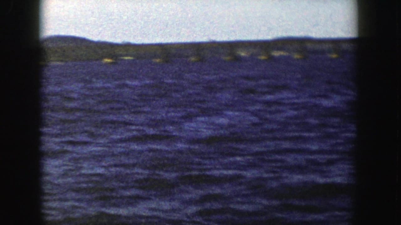 1958 Wisconsin 8mm frame showing open rippled water, a faint long bridge and low shoreline beneath a pale sky.