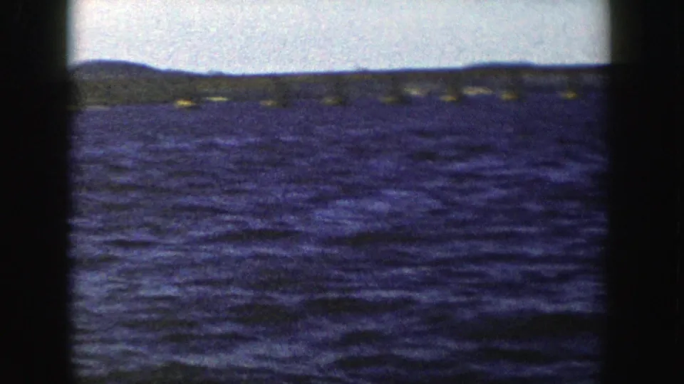 1958 Wisconsin 8mm frame showing open rippled water, a faint long bridge and low shoreline beneath a pale sky.