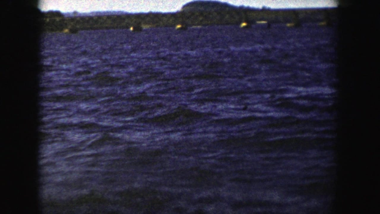 1958 Wisconsin 8mm frame looking across blue water toward a low bridge and dark shoreline, with the vessel no longer prominent.