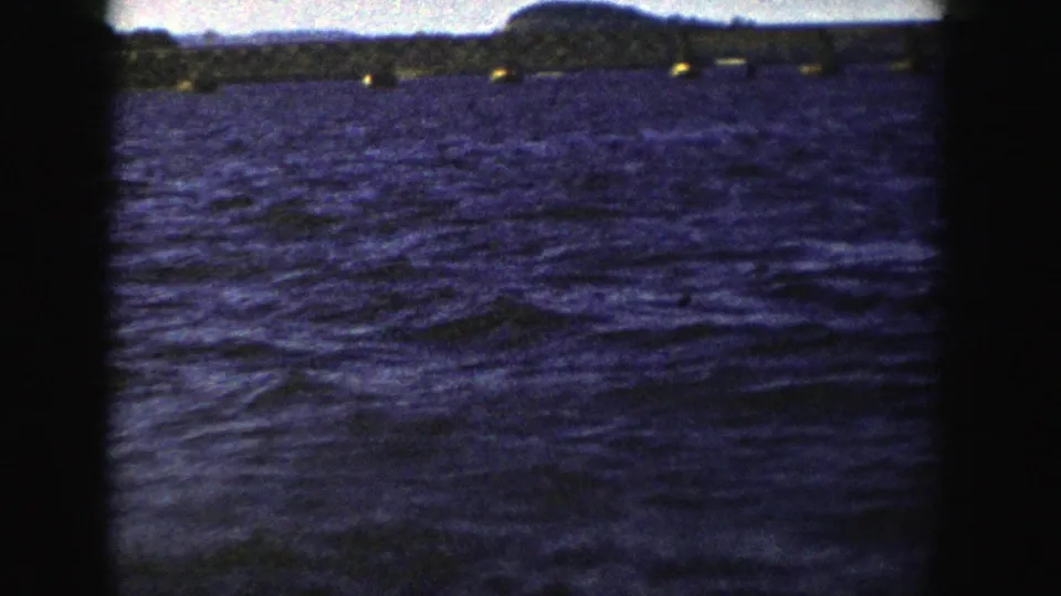 1958 Wisconsin 8mm frame looking across blue water toward a low bridge and dark shoreline, with the vessel no longer prominent.