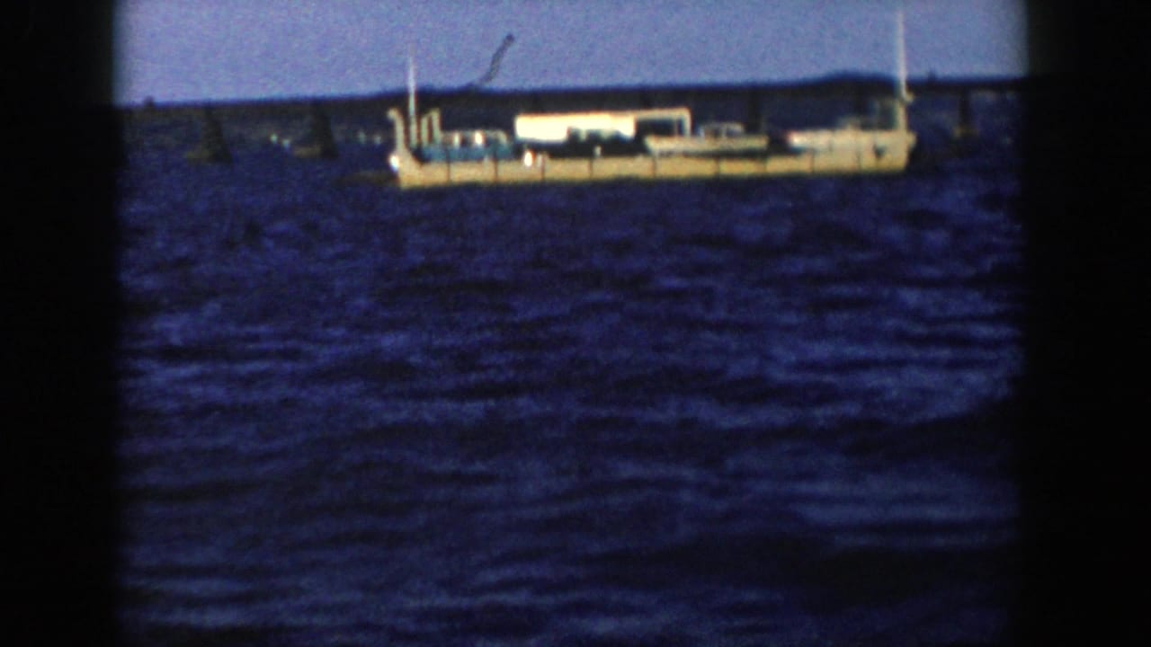 The tan vessel is framed closer on blue water, with soft 8mm color and a low Wisconsin shoreline in the distance.