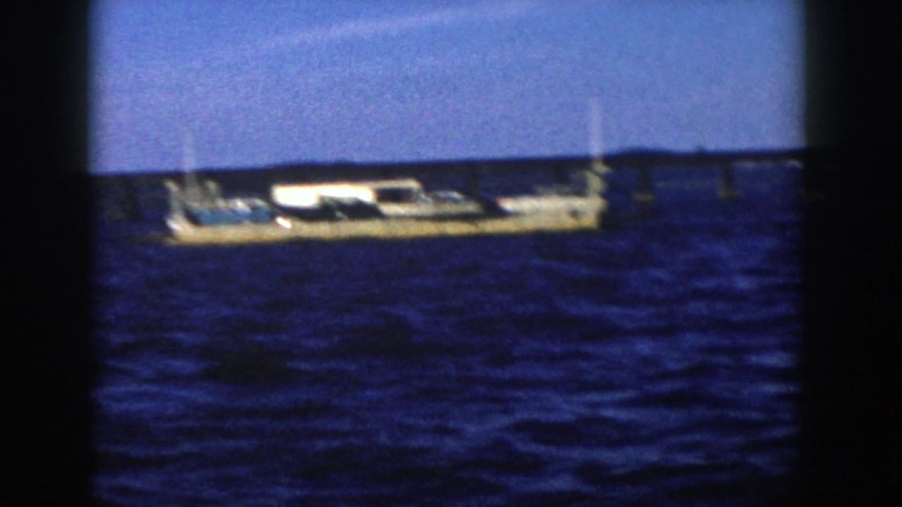 A long tan barge-like vessel moves across blue water in a grainy 1958 Wisconsin home-movie frame.