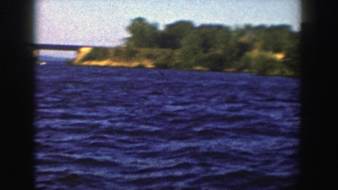 Blue lake water seen through dark boat or window framing with wooded shoreline and a small bridge in the distance.