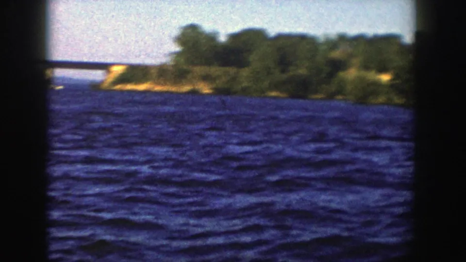 1958 Wisconsin 8mm frame showing rippled blue lake water, dark vertical framing, wooded shore, and a distant low bridge.